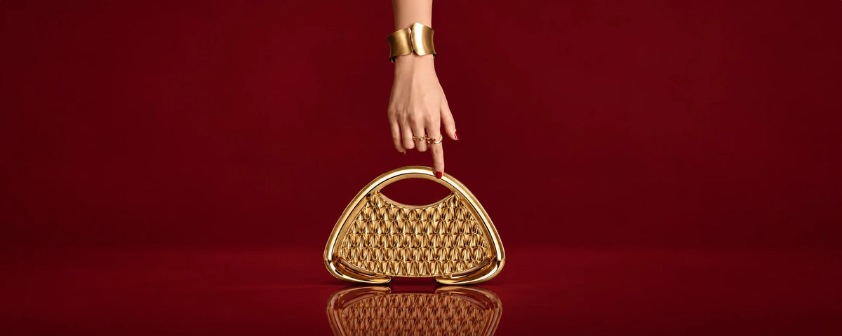 Gold sculptural handbag held by a hand with a gold cuff against a deep burgundy backdrop